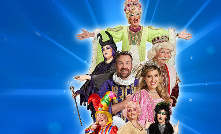 All eight main cast members are gathered in the centre of the image on a blue gradient and radiating light from behind and with a few sparkles. At the top of the image is Our Dame – John Furlong in a bright green dress with feathers and their arm outstrectched. Below them is Magnificent – Crystal Lee Murphy dressed in black and purple on the left and King Anto – John Cunningham to the right in a crown and royal coat. Below them close together is Prince William – Cormac Dredge wearing a crown and a royal purple coat and Aurora – Ailbhe Travers, in a crown and pink ballgown. At the bottom is Tickles – Dermot Byrne on the left in a red colourful Jesters costume, in the centre The Fairy – Aoife Dixon wearing a golden dress, with pink hair and fair wings and then on the right Rubella – Amy Ward, wearing green and black with their hand held up to their shoulders on either side doing the 'rock on' gesture.